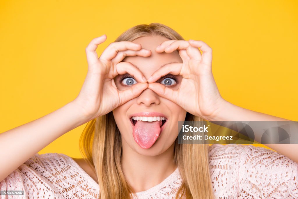 Head shot close up portrait of foolish playful girl gesturing tongue-out making binoculars with ok signs looking at camera isolated on yellow background Women Stock Photo