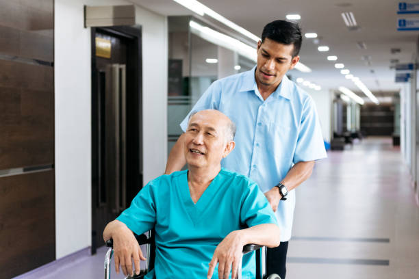 Senior man being pushed in wheelchair by male nurse Malaysian man in his 70s smiling and looking away, male porter pushing him along hospital corridor male porter stock pictures, royalty-free photos & images