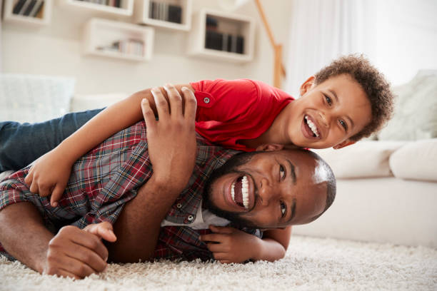 Son Climbs On Fathers Back As They Play Game In Lounge Together Son Climbs On Fathers Back As They Play Game In Lounge Together african father stock pictures, royalty-free photos & images