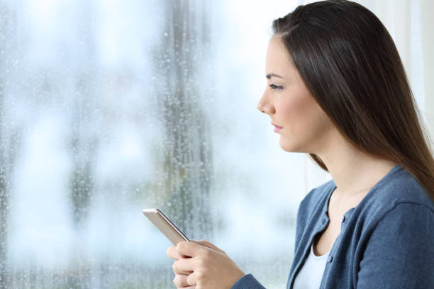 istock Sad woman holding phone and looking through a window 976969806