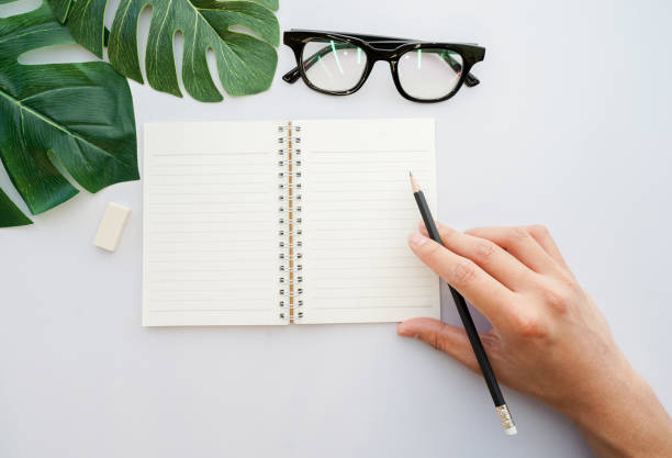 istock close up man hand using black pencil and prepared to writing on mock up notebook in top view with tropical leaves , rubber and glasses on white table background for lifestyle concept 977152560