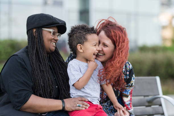 Two mothers holding their son on their laps outside at the park A homosexual biracial couple sit on a park bench with their young son. He is sitting on their laps and they're snuggling and talking with him. One mom is African-American and the other is Caucasian. surrogacy stock pictures, royalty-free photos & images