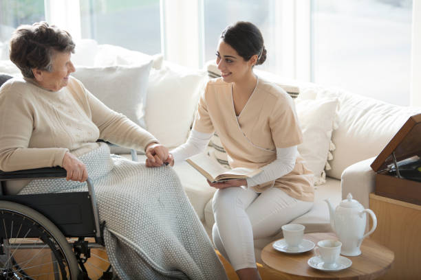 Disabled woman with medical assistant A disabled old woman on a wheelchair holding hand of a tender professional medical assistant during tea time in a living room of luxury retirement home military private stock pictures, royalty-free photos & images