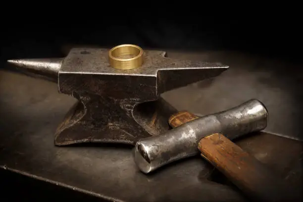 golden ring on an anvil and a goldsmith hammer in the jewelry workshop, still life with copy space in the dark background, selected focus, narrow depth of field