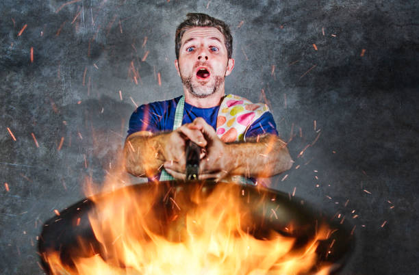 young attractive and shocked messy home cook man with apron holding pan in fire burning the food in kitchen disaster and unskilled and unexperienced terrible home cook at domestic cooking hombre joven atractivo y sorprendido sucio cocinero del hogar con delantal sosteniendo la sartén en el fuego que quema el alimento en el desastre de cocina y cocinero del hogar terrible no calificado y sin experiencia en la cocina doméstica