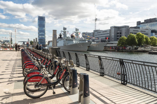 Santander sponsored London bicycles at Canary Wharf in London Also visible are two Chinese Warships, The Huanggang and Yangzhou, which docked at Canary Wharf as part of a goodwill tour bicycle docking station stock pictures, royalty-free photos & images