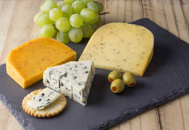 istock Cheese board with three cheeses, gouda with pimento, gouda with cumin seeds and roquefort blue cheese close up on rustic wooden background 977670192
