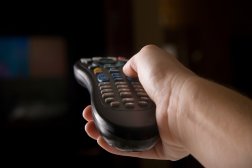 A hand pointing the remote towards the hint of a television in a dark background.
