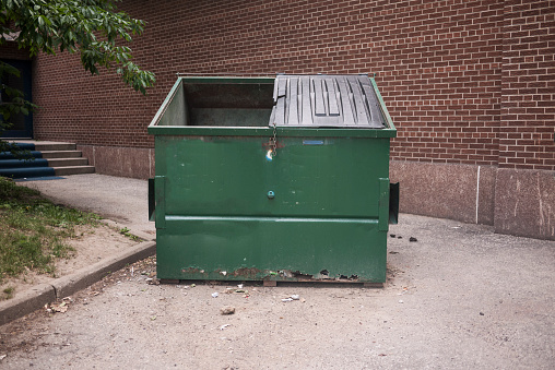 Damaged dark green dumpster near a brick building.