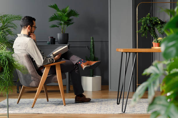 современный человек читает книгу, сидя в сером кре�сле в модном интерьере с растениями - relaxation indoors reading one person стоковые фото и изображения