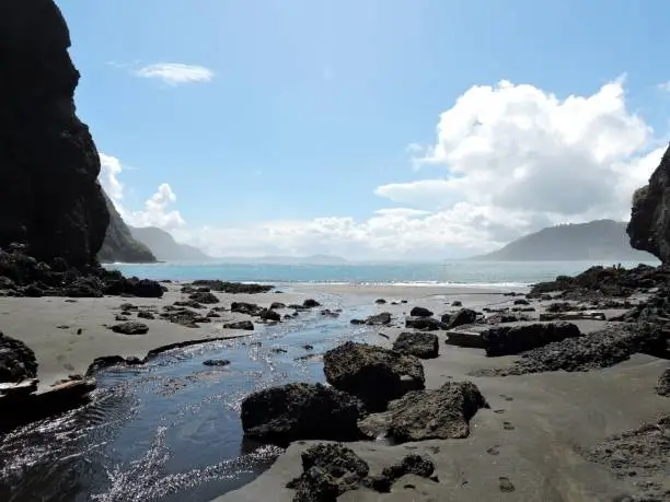 On a lonely black beach, a river / tide-way flows into the sea, decorated by rocks, New Zealand