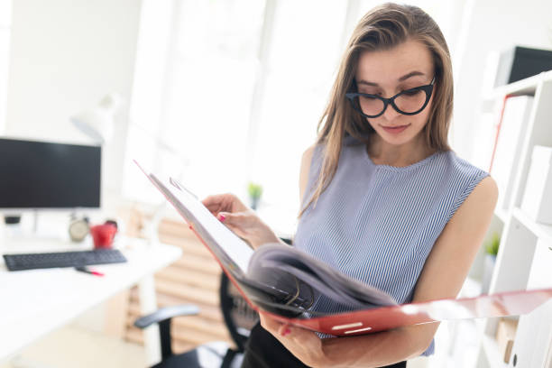 istock Beautiful young girl in an office stands near a rack and scrolls through a folder with documents. 977976468