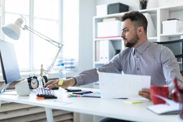 istock A young man in the office sits at a table, holds a marker in his hand and works with documents and a computer. 977976544