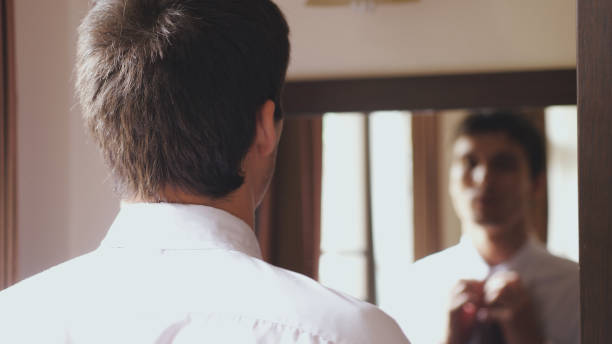 istock Man in white shirt stands by the mirror tying a tie 978149170