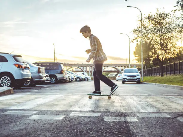 Photo of young hipster boy crossing the street riding the skateboard in the urban city