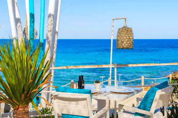 Photo of White table and chairs with blue pillows on restaurant terrace on Cala Nova beach with beautiful sea view, Ibiza island, Spain