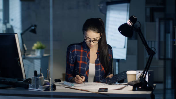 istock Late at Night Female Solar Engineer Works on Drafts, Holds Part of Solar Panel and Uses Her Personal Computer. Office is Empty, Only Her Table Lamp and Moonlight Illuminates Room. 978339168