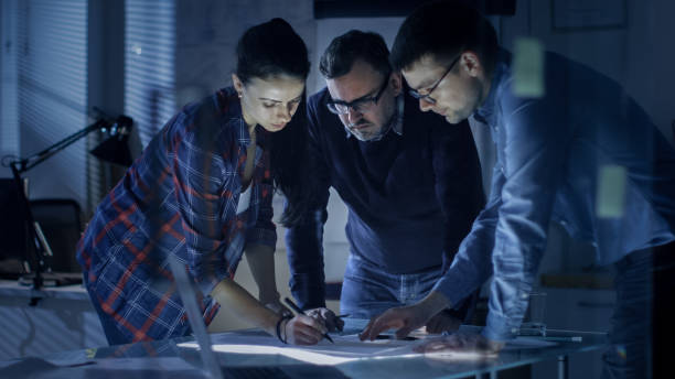 istock Late at Night Team of Design Engineers Collectively Work on the Project. They Inspect Drafts, Documents and Have Discussion at the Big Table. 978339194