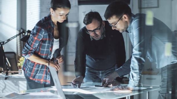 istock Late at Night Team of Design Engineers Work on a Project at the Big Conference Table. They Exchange Opinions and Make Corrections in Documents. Table is Illuminated. 978339200