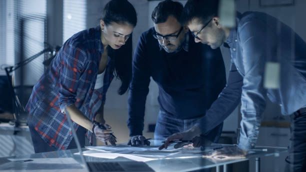 istock Late at Night Team of Design Engineers Collectively Work on the Project. They Inspect Drafts, Documents and Have Discussion at the Big Table. 978339202