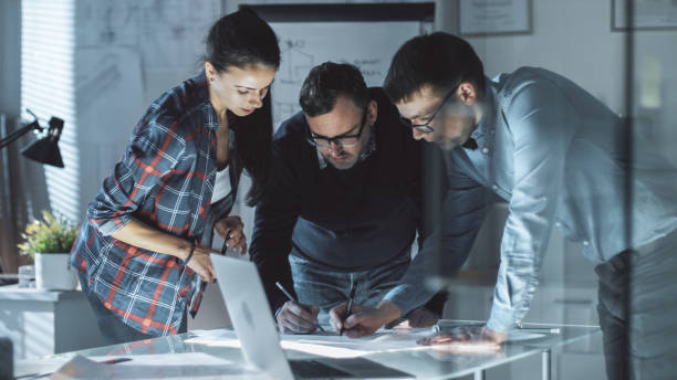 istock Late at Night Team of Design Engineers Work on a Project at the Big Conference Table. They Exchange Opinions and Make Corrections in Documents. Table is Illuminated. 978339206