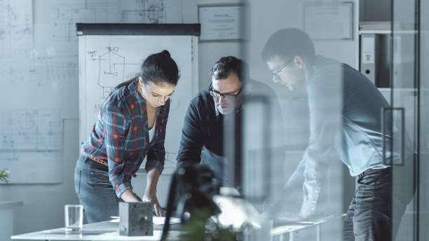 istock Team of Design Engineers Work on a Project. They Work on a Illuminated Conference Table with Documents, Blueprints and Laptop on it. 978339228