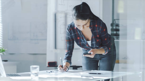 istock Female Design Engineer Works on Documents in a Conference Room, Last Minute Check-up, Uses Her Smartphone. In the Background Whiteboard with Schemes on it, Various Blueprints Hanging on the Walls. 978339262