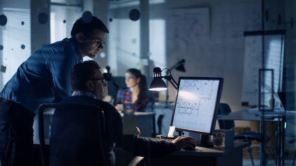 istock Late at Night Two Men Discuss Blueprints that are Shown on Display of a Personal Computer. Office Looks Modern with Lots of Blueprints on the Walls. In the Background Woman Working. 978339322