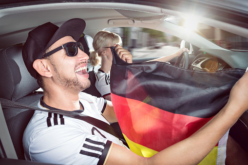 A male soccer fan inside a car is holding a German flag. He is celebrating a win. In the background a female is driving the car.