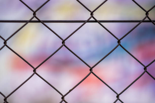 An abstract patterend shot of wire mesh fencing with blurred colorful graffiti in the background in Johannesburg inner city This fence cordoned off an area, behind which there are walls covered in graffiti. The ability to access inaccessible areas is a key skill in graffiti culture purple graffiti metallic wall stock pictures, royalty-free photos & images