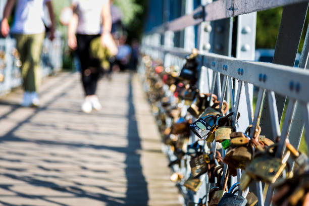 Close up color image depicting a collection of padlocks attached to the side of a footbridge. The padlocks have different messages on them, mainly declarations of love. In the background, defocused, people are walking across the bridge, which is in Cluj Napoca, in the Transylvania region of Romania. Room for copy space.