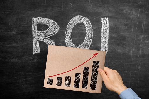 Man holding a hand drawn success graph chart on a cardboard over the word ROI (Return on Investment) written with chalk on blackboard