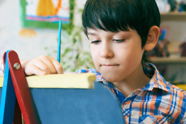 istock Child standingnext to the easel. Kid boy learn paint by brush in class school. Kindergarten interior on background. Boy is getting ready to become an artist 978415892