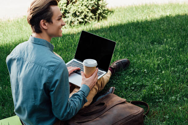 istock smiling young freelancer holding paper cup and using laptop with blank screen while sitting on grass 978462114