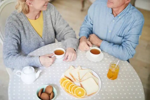 Overview of slices of bread on plate, jam in jar, fresh oranges and cookies and senior couple sitting by table and having tea