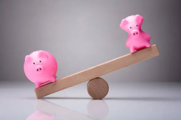 Unbalance Of Two Piggybanks On Wooden Seesaw Against Gray Background