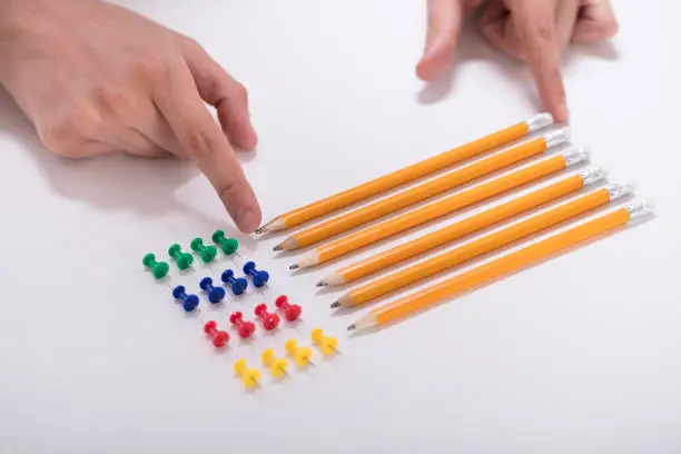 A Person's Hand Arranging Pencils And Multi Colored Pushpins In A Row On White Background