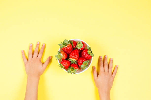 istock Kid hands and bowl with fresh strawberries on yellow background. 978573976