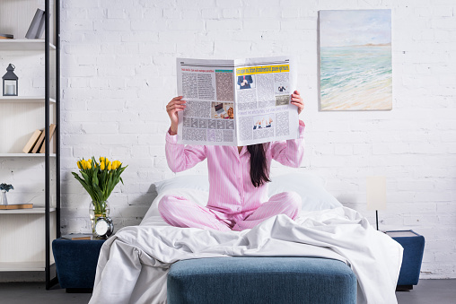 obscured view of woman in pajamas with newspaper in bed at home