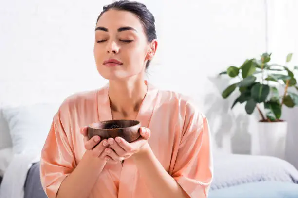 Photo of woman in silk bathrobe with wooden bowl in hands having tea ceremony in morning at home
