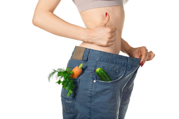Weight loss and healthy eating or dieting concept. Slim girl in oversized jeans with raw vegetables in the pockets is holding her thumb up. Side view. Close shot stock photo