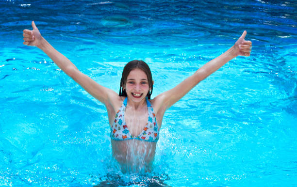 Slim young girl smiling at the camera is spectacularly jumping out of pool full of clear blue water