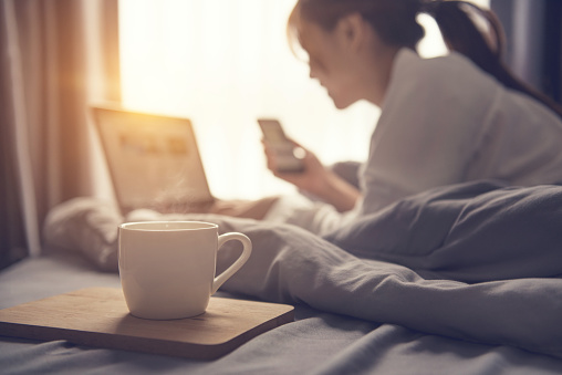 Close up coffee cup and blurred Woman wake up and working with laptop and smartphone start up project on bed in bedroom, Early morning