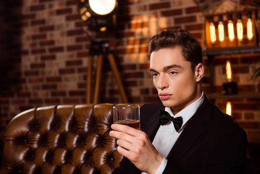 Vertical portrait of virile perfect stunning man in black suit with bow-tie holding glass with cognac in hand looking to the side over brown brick wall, sitting on leather sofa