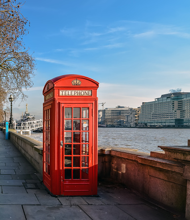 Classic British red telephone box at Thames embankment in London center. This telephone kiosk is a familiar sight on the streets of the United Kingdom. The paint colour used today for the boxes is known as \
