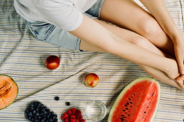 summer picnic setting. Woman in jean shorts and hat eatining water melon, melon, blueberries, raspberries and peaches, drinking water. Outdoor gathering concept. stock photo