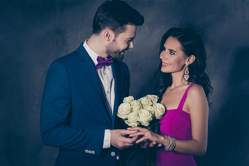 Portrait of trendy gentleman in tux with bow putting on gold diamond ring with diamond on finger to joyful cheerful lady with jewelry holding white roses in hands, couple isolated on grey background