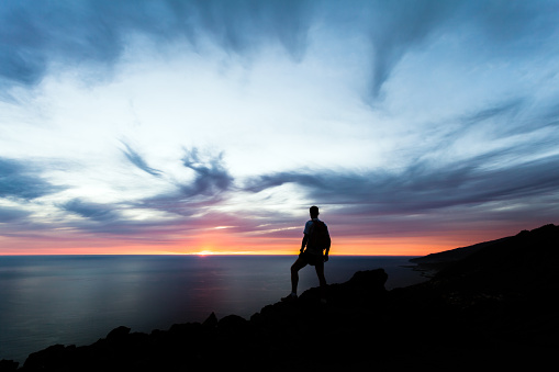 Celebrating or meditating man looking at sunset ocean. Hiker with backpack on top of mountain looking at beautiful inspirational night landscape.