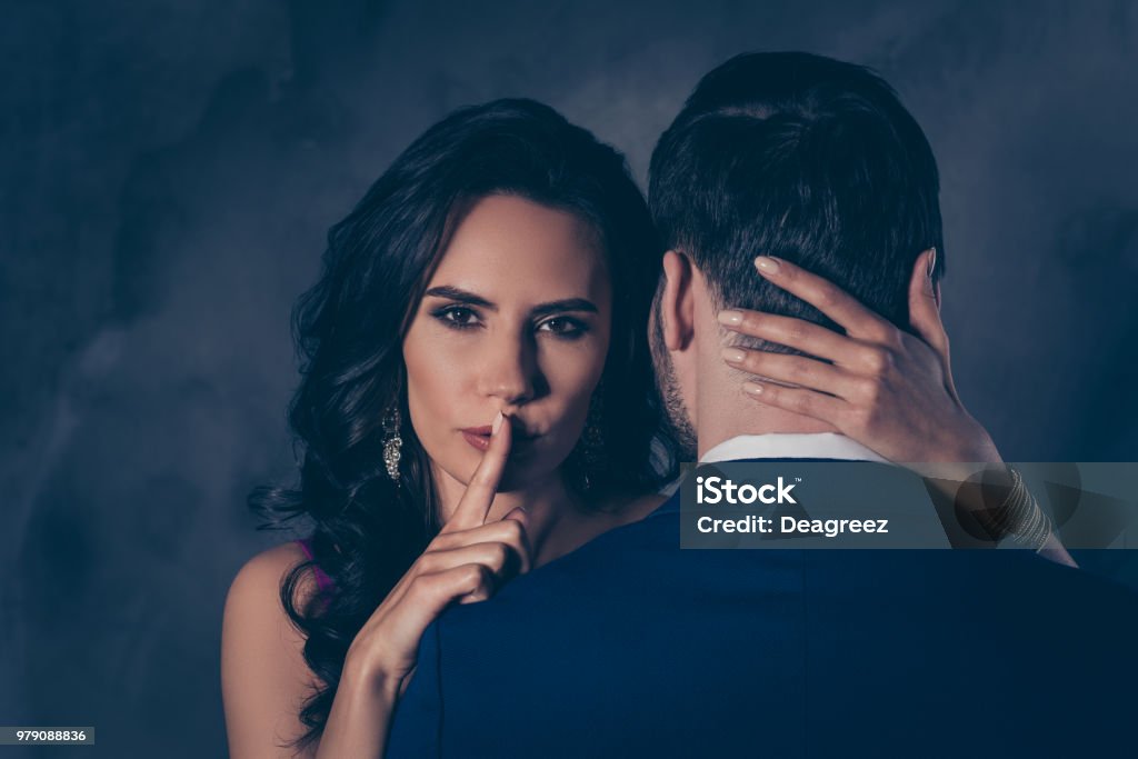 Shh! Portrait of tempting brunette lady showing silence sign with forefinger touching secret mysterious gentlemen with rear view, lovely Mr and Mrs isolated on grey background Infidelity Stock Photo