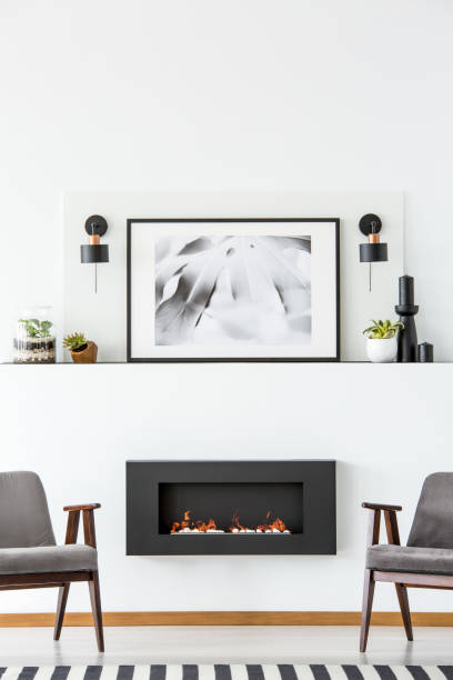 istock Black fireplace between grey armchairs in white flat interior with poster between lamps. Real photo 979189682